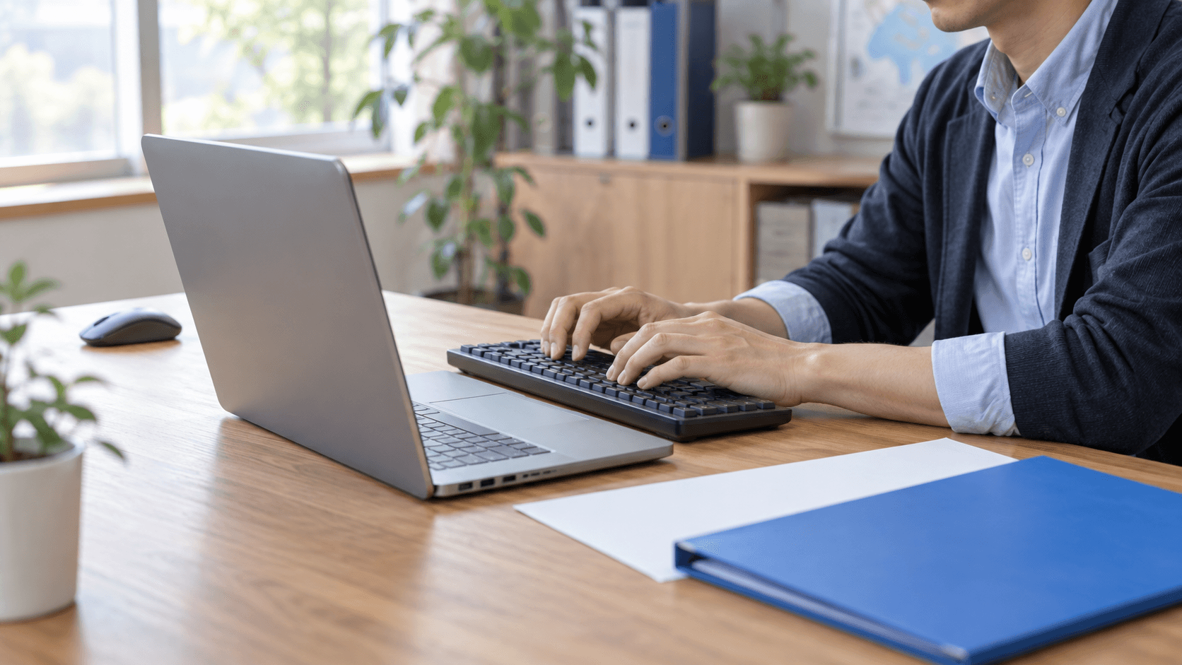明るい事務所で担当者がノートPCにつないだ外付けキーボードを操作している。マウスは手元から離れ、机には青いフォルダーと白い用紙が置かれている