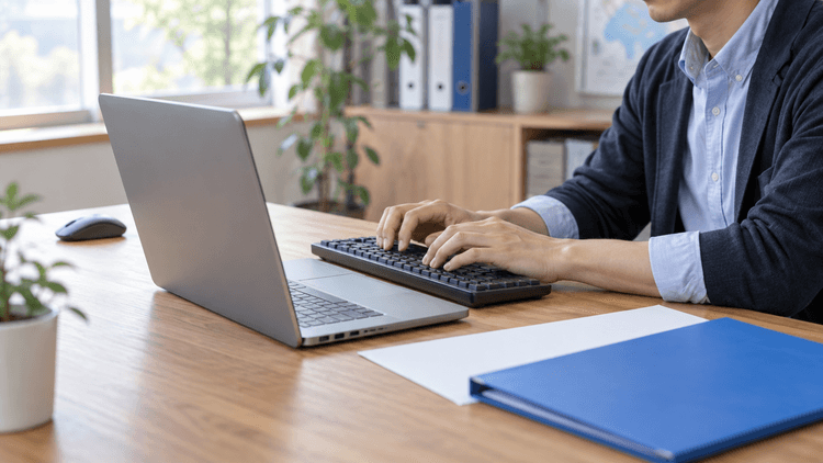 明るい事務所で担当者がノートPCにつないだ外付けキーボードを操作している。マウスは手元から離れ、机には青いフォルダーと白い用紙が置かれている