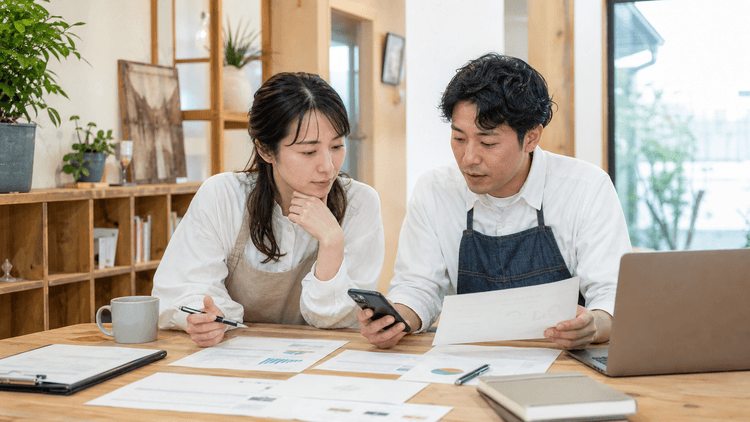 地域の店舗担当者と広報担当者が机の上に案内資料とスマートフォンを広げ、店舗情報とよくある質問の整理方針を打ち合わせている様子
